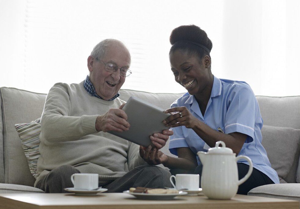 Happy senior man and nurse using digital tablet at home - Cahoon Care Associates Home caregiver and elderly man laughing together